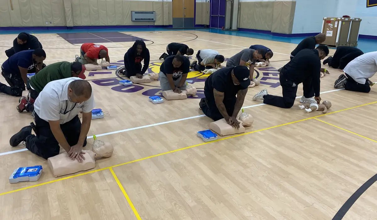 A group of adults is training in CPR in a basketball gym.