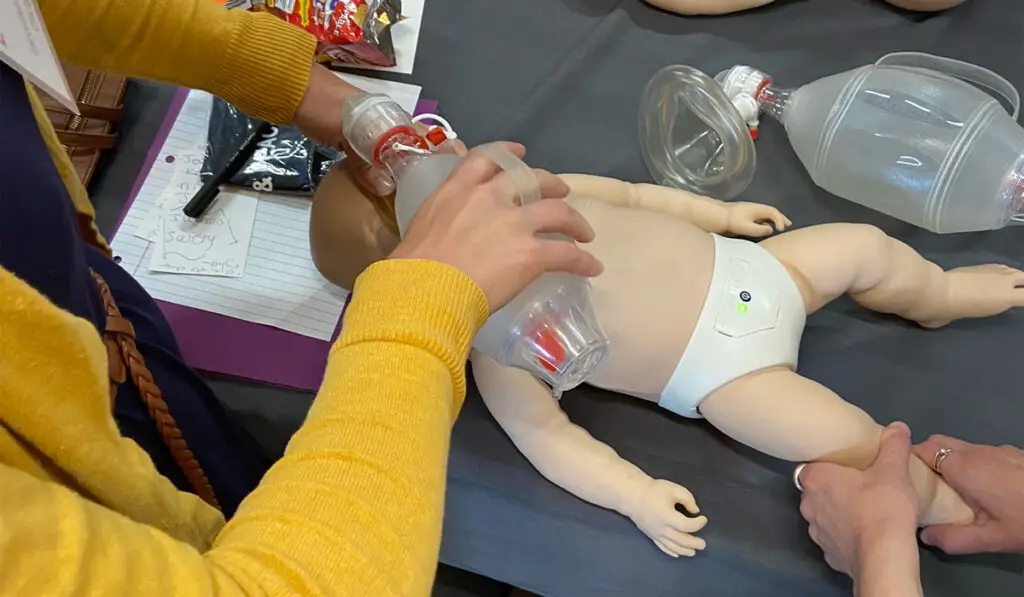 Hands manually resuscitating a baby dummy using a bag valve mask.