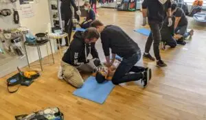 Two trainers demonstrating advanced CPR techniques on a high-tech mannequin in a training workshop filled with medical equipment.