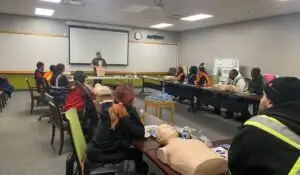 CPR Certification training session in a classroom setting with participants seated at tables, practicing on mannequins while an instructor demonstrates.