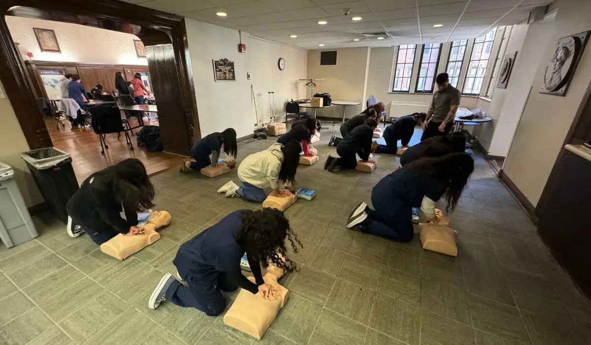 Multiple trainees performing CPR Certification training on dummies spread out on the floor in a spacious room with a trainer supervising.