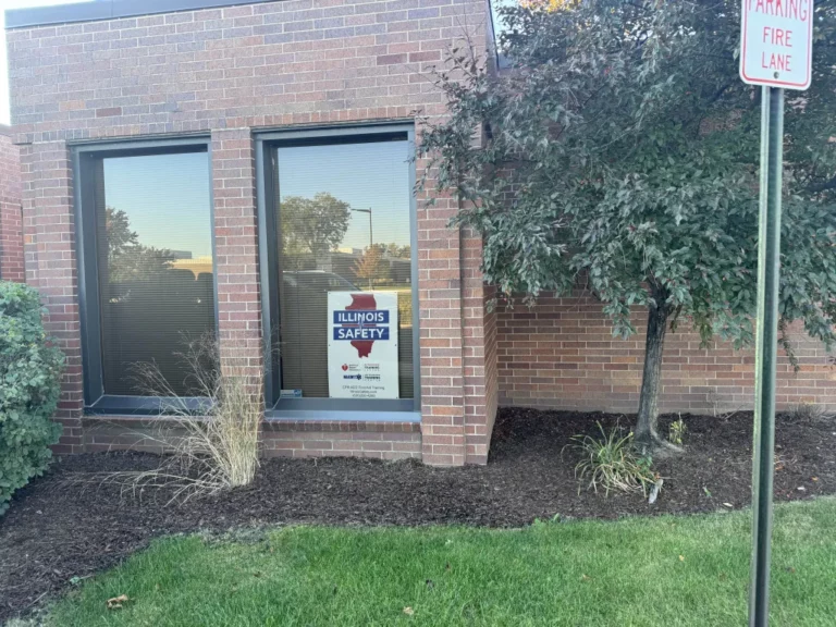 Exterior window of a brick building displaying a sign for Illinois Safety CPR-AED-First Aid Training, with a small tree and landscaping.